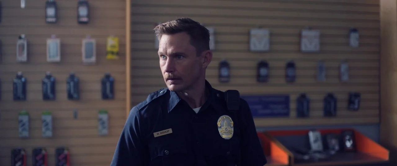 Blindfire (2020) movie still: medium — A man in a police uniform stands in front of a retail slatwall display. Numerous small items are packaged and…