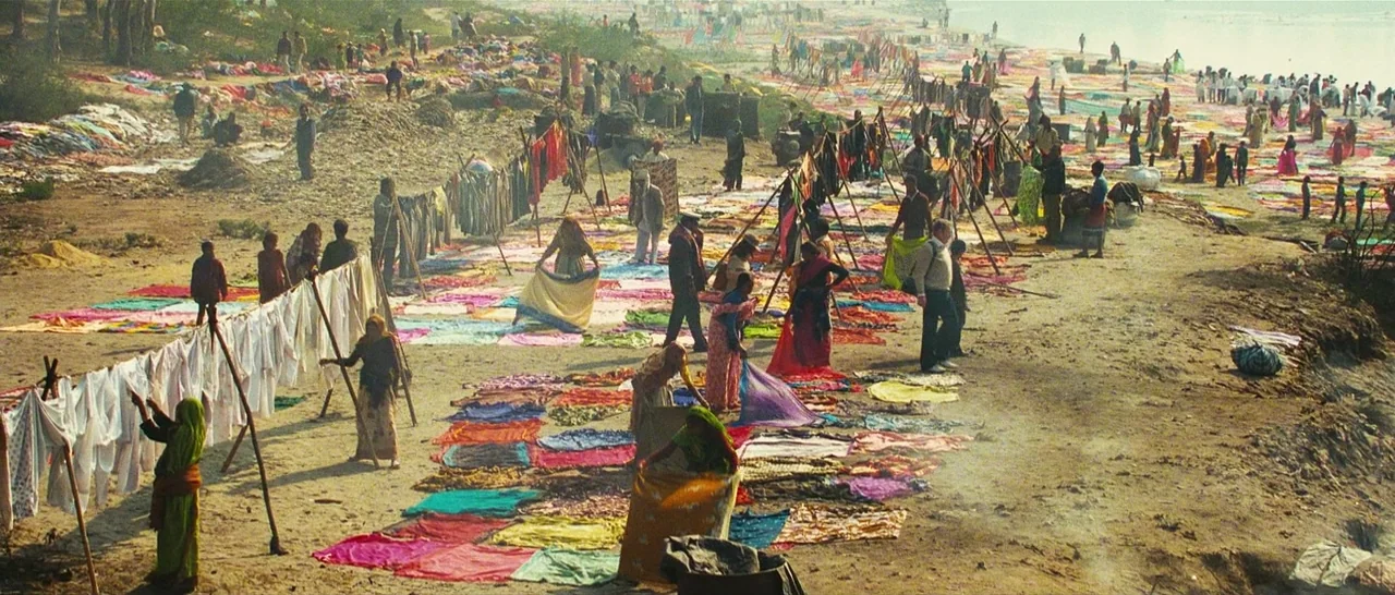 Slumdog Millionaire (2008) movie still: wide — A vast sandy riverbank is covered with rows of drying colorful fabrics and clothes. Numerous people…