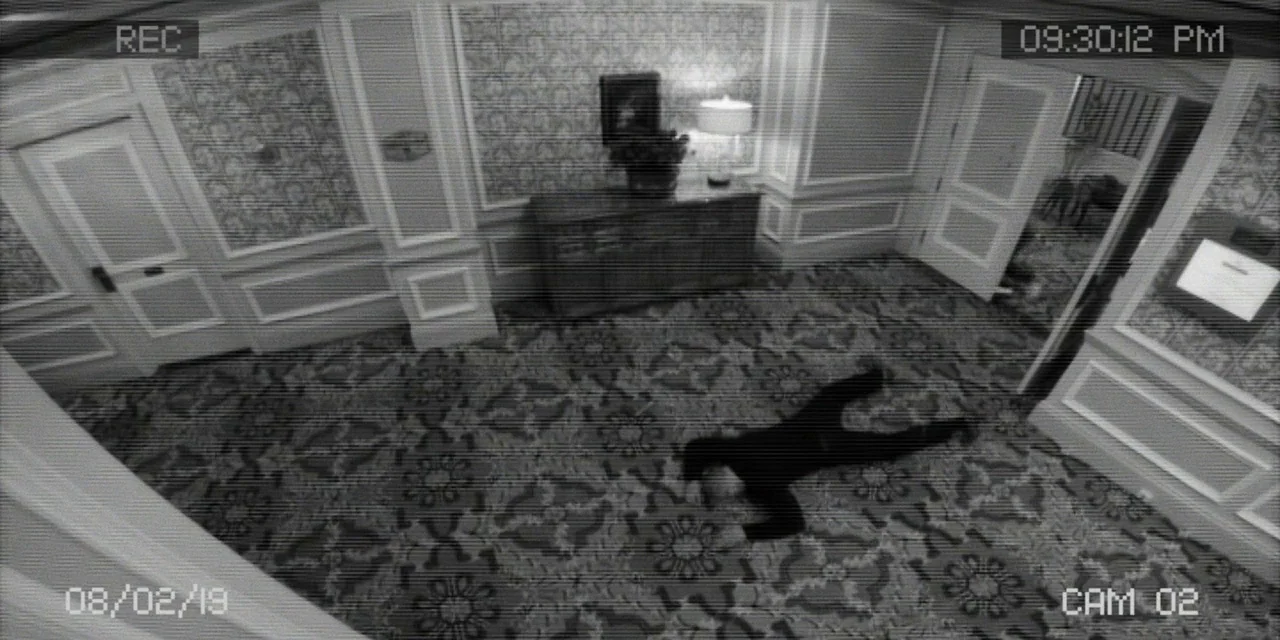 Polar (2019) movie still: wide — A high-angle surveillance view of a carpeted hotel hallway with patterned wallpaper and doors. A person in dark…