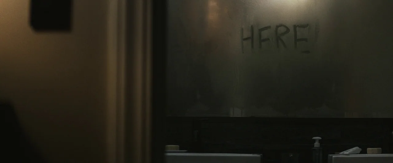 The Night House (2020) movie still: medium — A dimly lit bathroom mirror is covered in heavy condensation. The word HERE is written in the steam on…