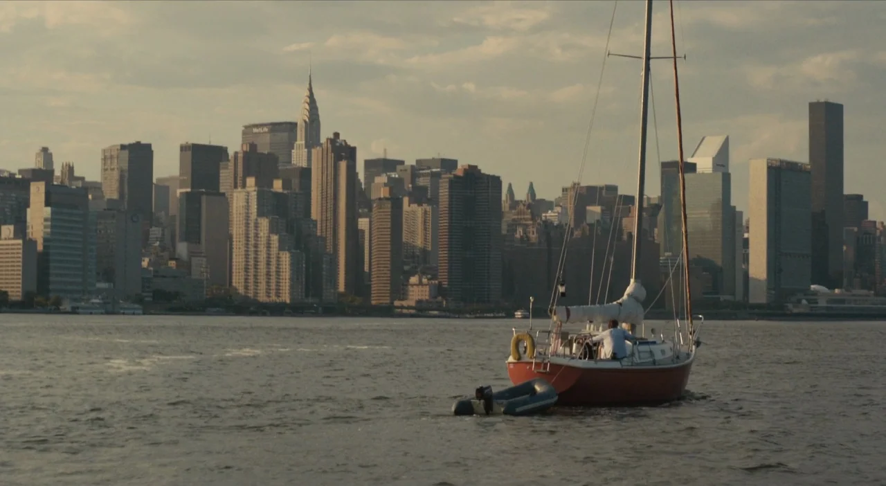 The Comedy (2012) movie still: wide — A sailboat moves across the East River with the Manhattan skyline in the distance. The urban architecture is…