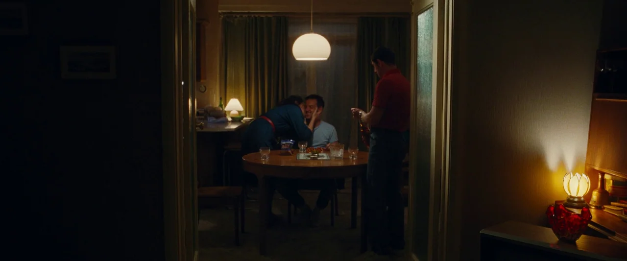 All Of Us Strangers (2023) movie still: wide — A dimly lit room contains a circular wooden table where three people are gathered. The woman leans in…