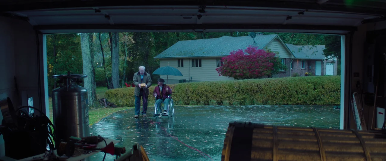 Linoleum (2022) movie still: wide — A wet driveway extends from a garage toward a suburban house with a hedge. An elderly man stands while another…