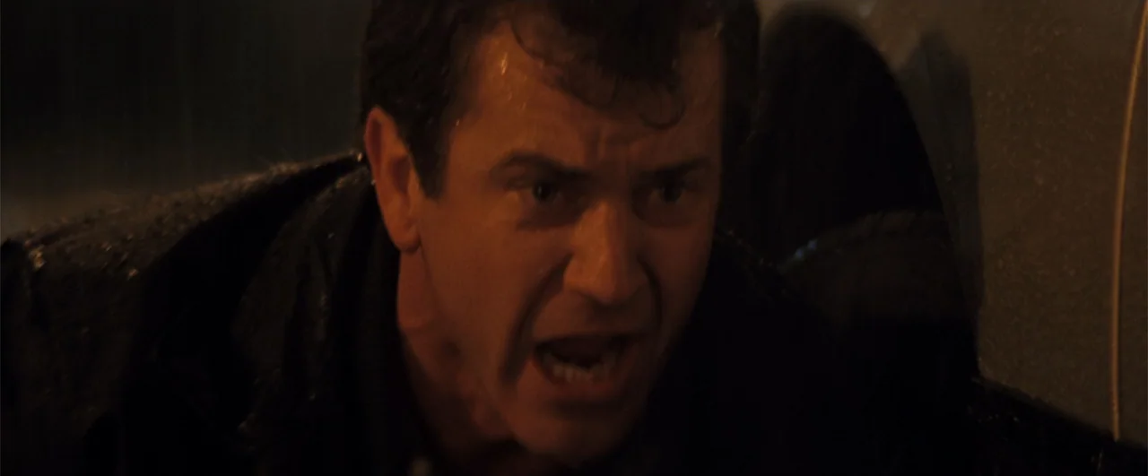Lethal Weapon 4 (1998) movie still: closeup — A dark, rain-slicked urban street at night with high-contrast lighting. A man with wet hair and a dark…