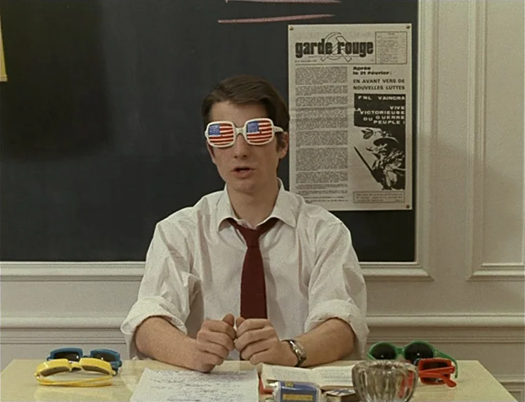 La Chinoise (1967) movie still: medium — A man sits at a desk in front of a black chalkboard with a poster. He wears novelty glasses with American…