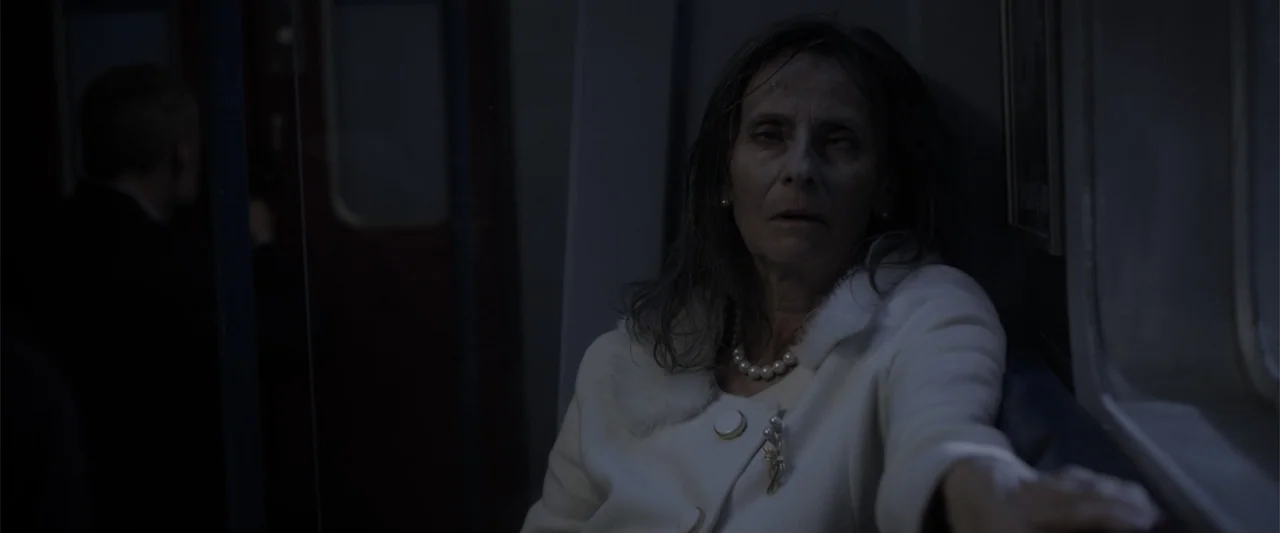 Howl (2015) movie still: medium — A dimly lit subway car interior with a single passenger on a bench. An elderly woman in a white coat sits staring…
