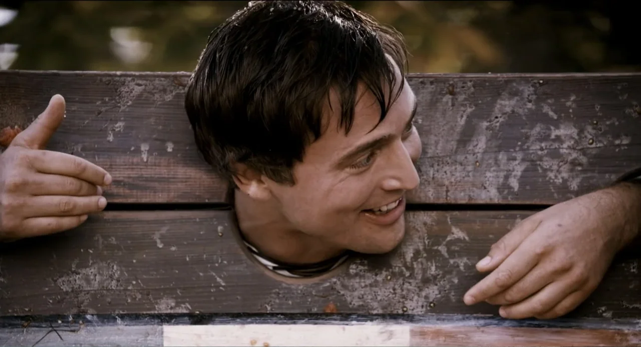 Todo es amor (2024) movie still: closeup — A man's head and hands are confined within a weathered wooden pillory structure. The subject looks toward…