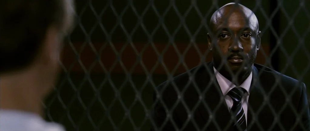 The Walker (2007) movie still: medium — A man in a suit sits behind a metal mesh divider in a room. The foreground shows the back of another…