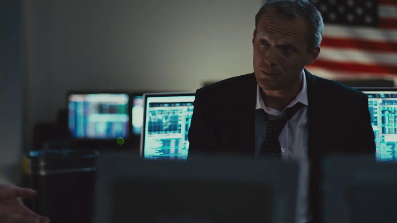 Margin Call (2011) movie still: medium — A dimly lit room filled with rows of computer monitors displaying complex stock market charts. A man in a…