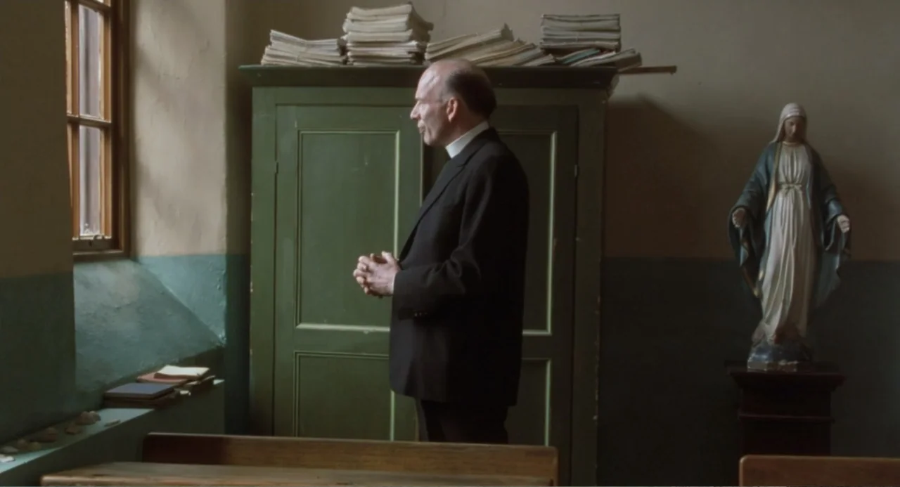 Dancing at Lughnasa (1998) movie still: medium — A spartan room features a green wooden cabinet with stacks of papers on top. A balding man in a…