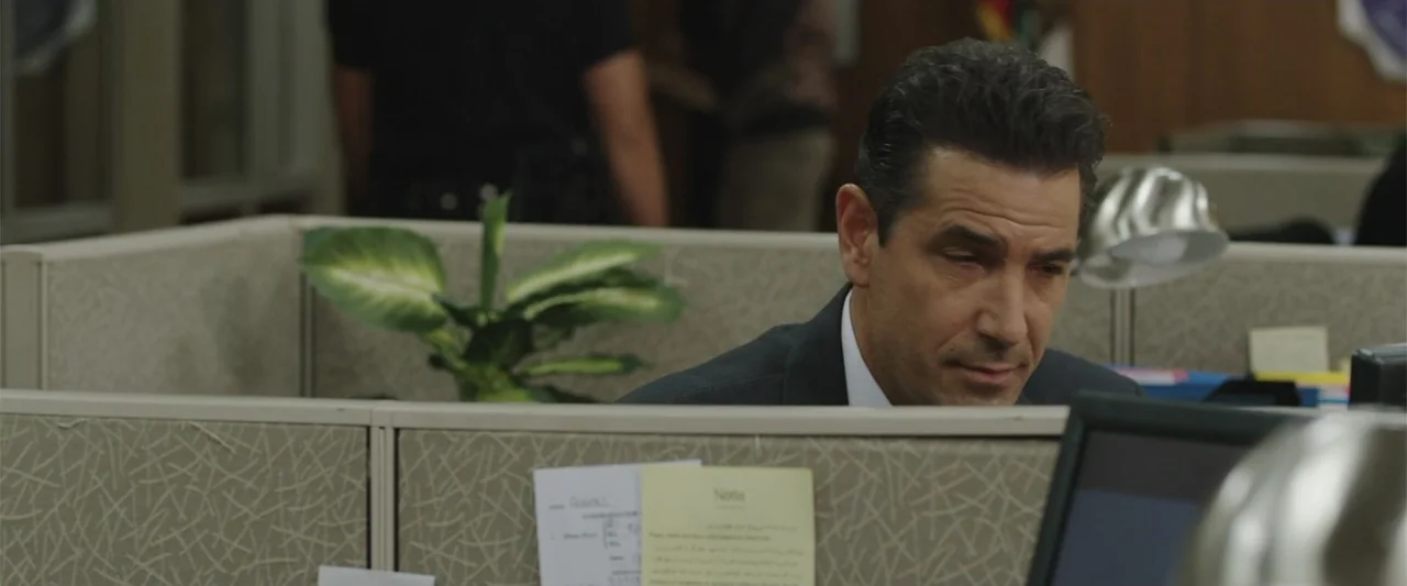 Presagios (2024) movie still: medium — A standard professional office space with grey fabric cubicle walls and a potted plant. A middle-aged man in…
