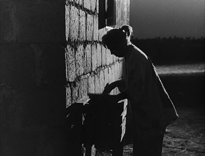Nights of Cabiria (1957) movie still: medium — A dark outdoor space features a rough-hewn stone wall next to a wooden workbench. A woman stands in…