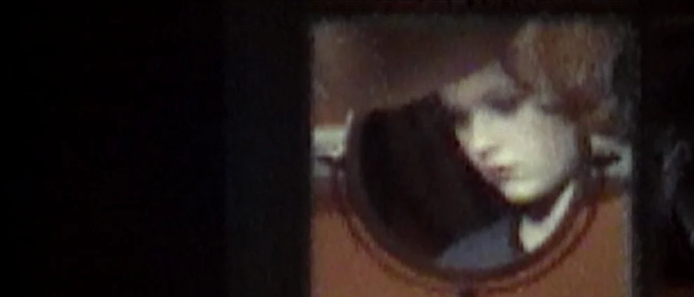 American Beauty (1999) movie still: closeup — A woman's face is reflected in a small, round mirror set against a dark background. She gazes downward…
