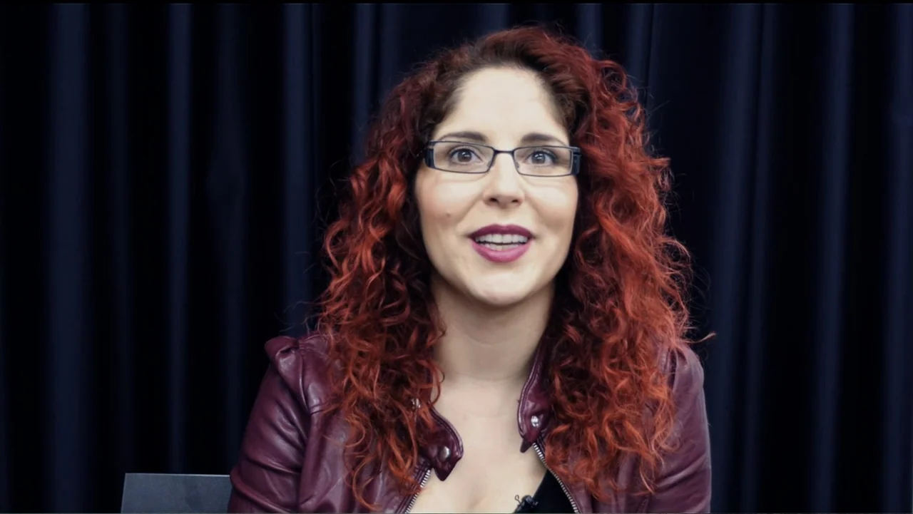 Soaring Highs and Brutal Lows: The Voices of Women in Metal (2015) movie still: medium — A woman stands in front of a dark blue curtain inside a…