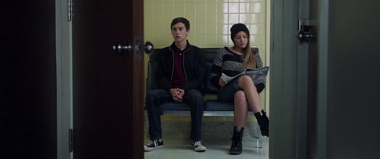 It Follows (2015) movie still: medium — A utilitarian waiting area features light yellow grid-patterned wall tiles and dark plastic seating. A young…