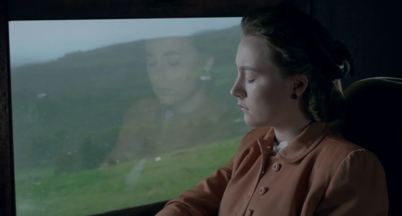 Brooklyn (2015) movie still: medium — A woman sits inside a train carriage looking out at the passing green landscape. Her reflection is cast upon…