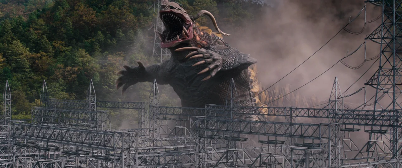 Shin Ultraman (2022) movie still: wide — A giant reptilian kaiju moves through a sprawling industrial electrical power substation. Clouds of dust…