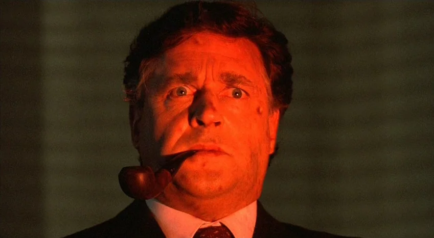The Coca-Cola Kid (1985) movie still: closeup — An interior office space is bathed in a distinct, concentrated red light. A middle-aged man stares…