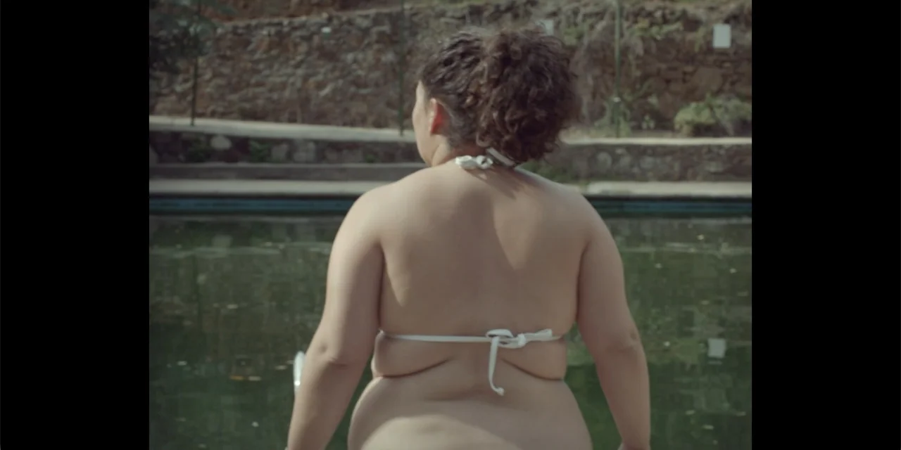 La coda del diavolo (2024) movie still: medium — An outdoor swimming pool with stone walls surrounds the water under daylight. A woman in a white…