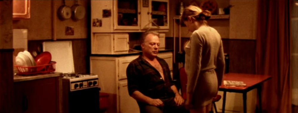 Carne (1991) movie still: medium — A dated 1990s kitchen with white cabinets, metal appliances, and a red table. A middle-aged man sits in a chair…