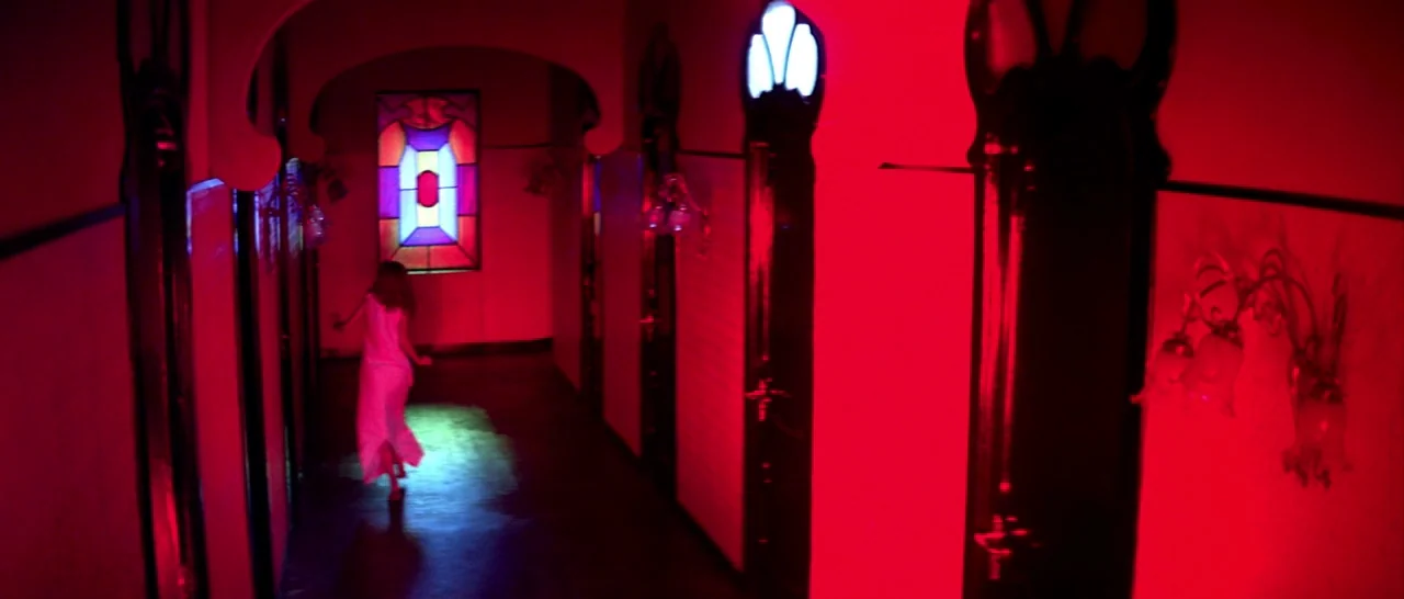 Suspiria (1977) movie still: wide — A long interior hallway features dark wooden paneling and a vibrant stained glass window. A woman in a white…