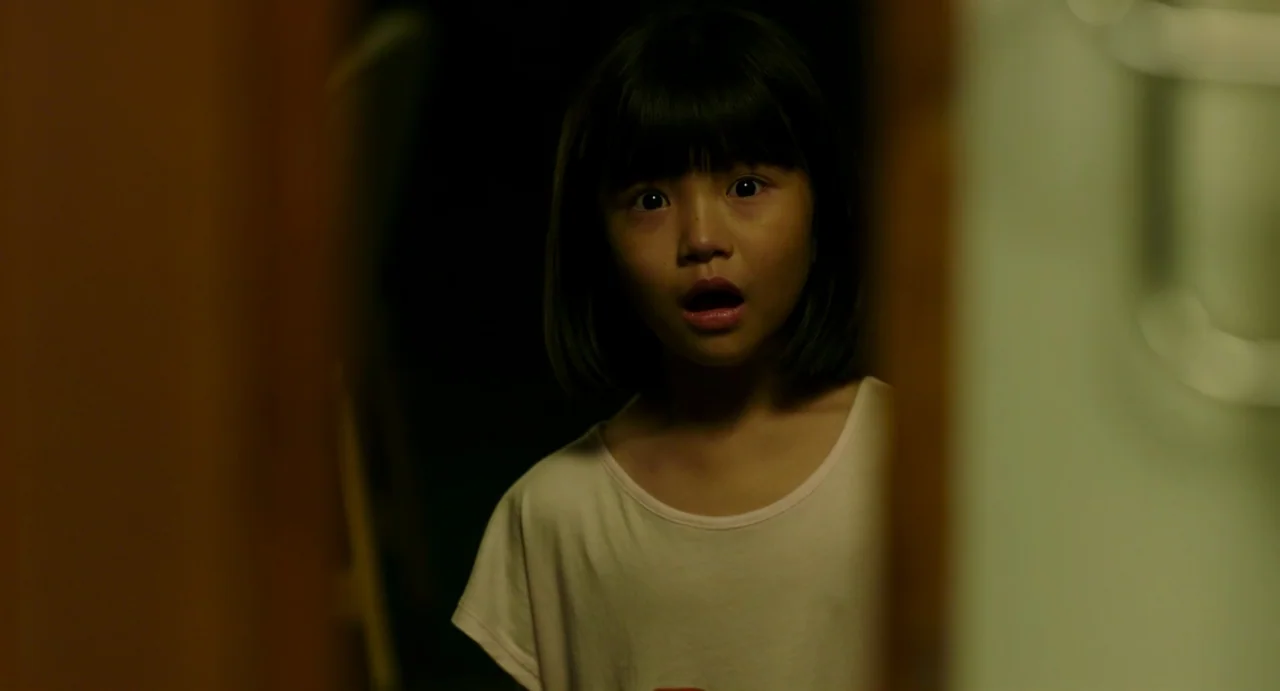 The Mimic (2017) movie still: closeup — A dark hallway transitions into a illuminated space where a child is framed partially. The child peers out…