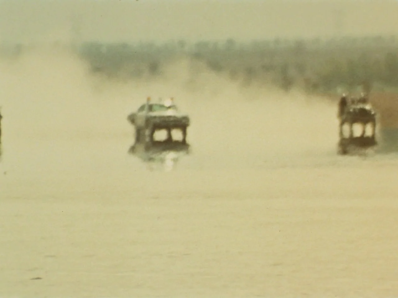 Punishment Park (1971) movie still: wide — A hazy, vast desert landscape is obscured by heavy heat shimmer and suspended dust. Two vehicles travel…