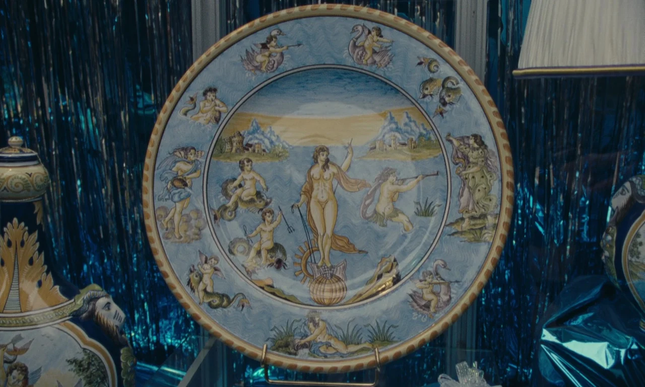 A Tale of Winter (1992) movie still: closeup — A decorative ceramic plate featuring classical figures is displayed against shimmering blue tinsel.…