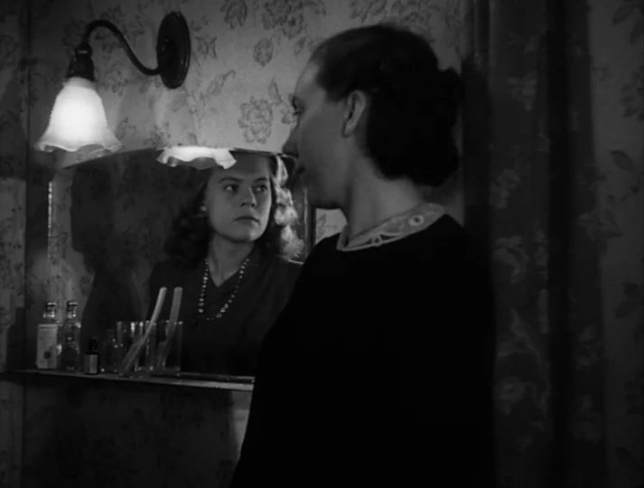 Port of Call (1948) movie still: medium — A small room features floral wallpaper and a mirror above a glass shelf. One woman stands in profile while…