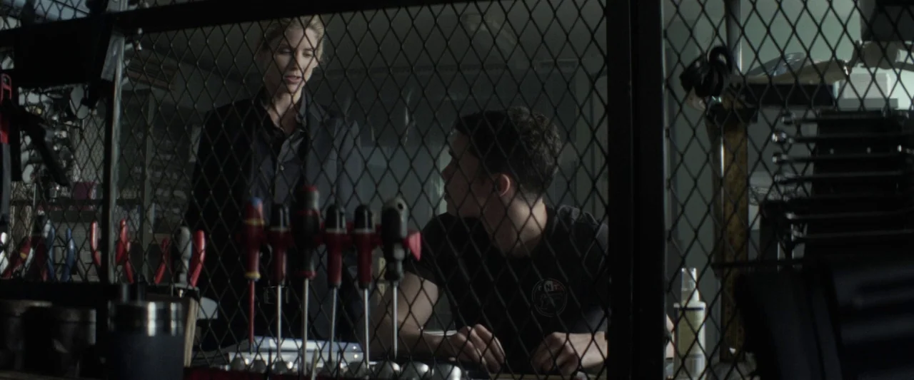 Colt 45 (2014) movie still: medium — A cluttered workspace featuring a metal chain-link fence barrier partitioning the foreground. A woman stands…