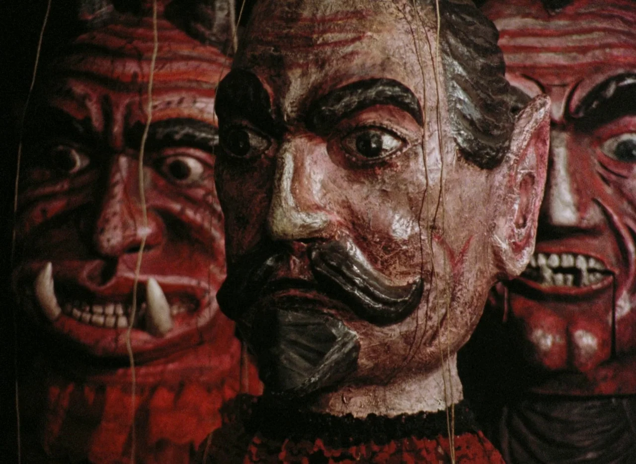 Faust (1994) movie still: closeup — Three wooden marionettes with red painted faces hang in a dark, interior space. Each puppet features hand-carved…