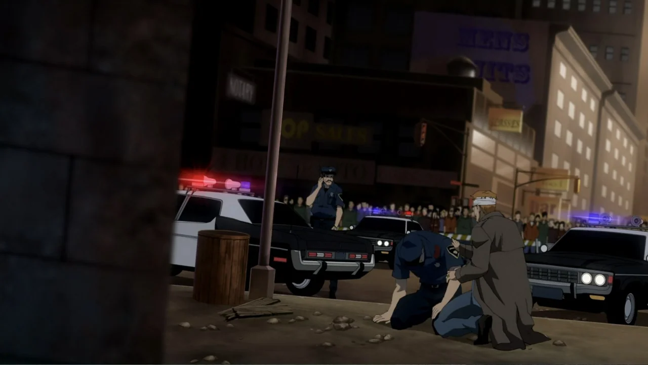 Batman: Year One (2011) movie still: medium — An urban city street at night featuring police cars with active blue and red lights. A man kneels on…