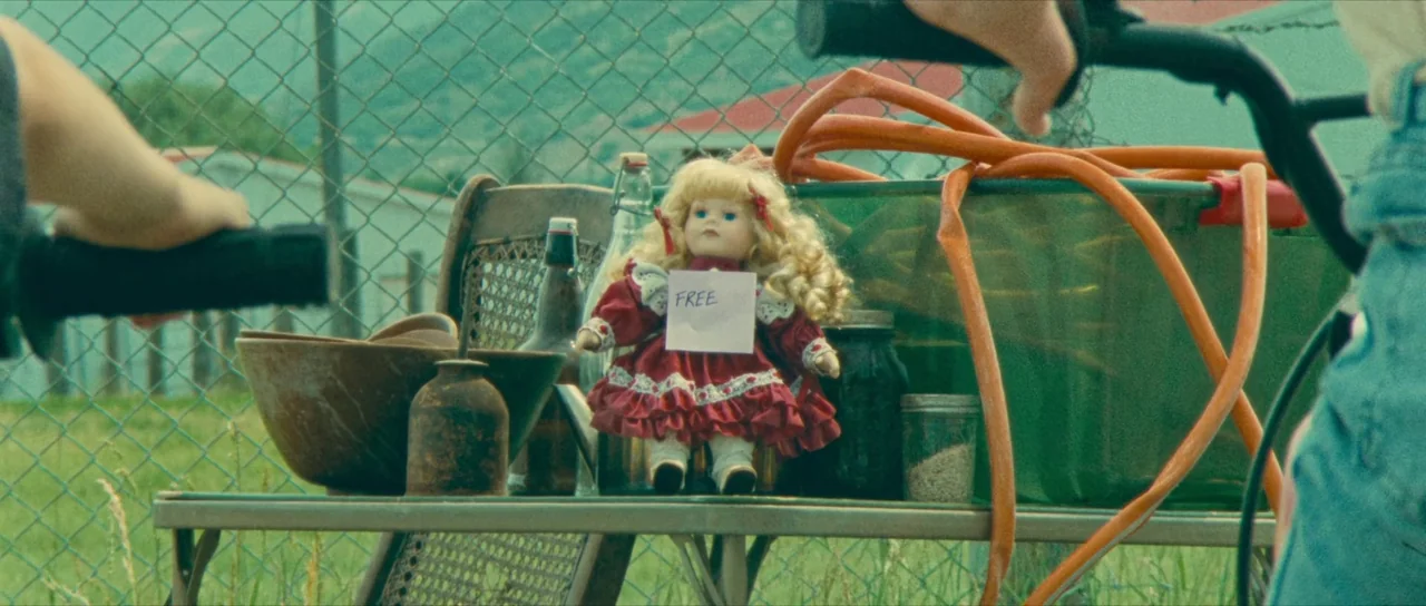 Riddle of Fire (2023) movie still: medium — A porcelain doll in a red dress sits on a metal table outdoors. A sign reading FREE is attached to the…