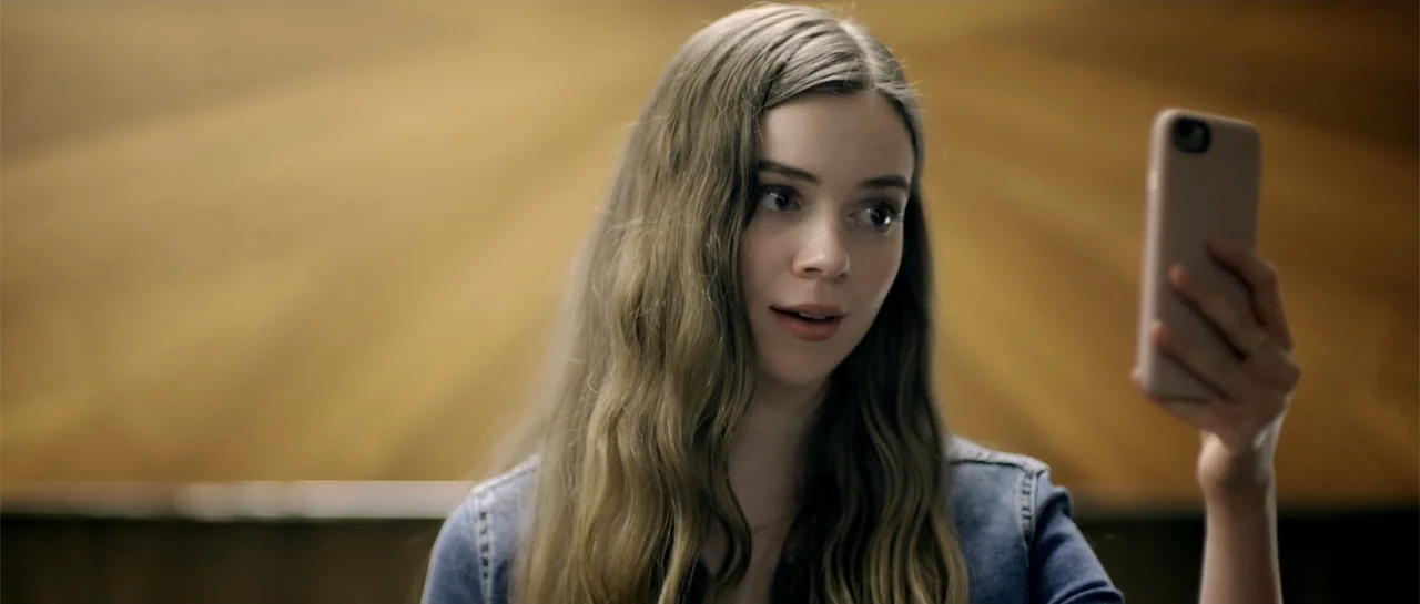 Elevator Game (2023) movie still: medium — A woman stands indoors in front of a warm-toned wooden paneled surface. She holds a smartphone up at eye…