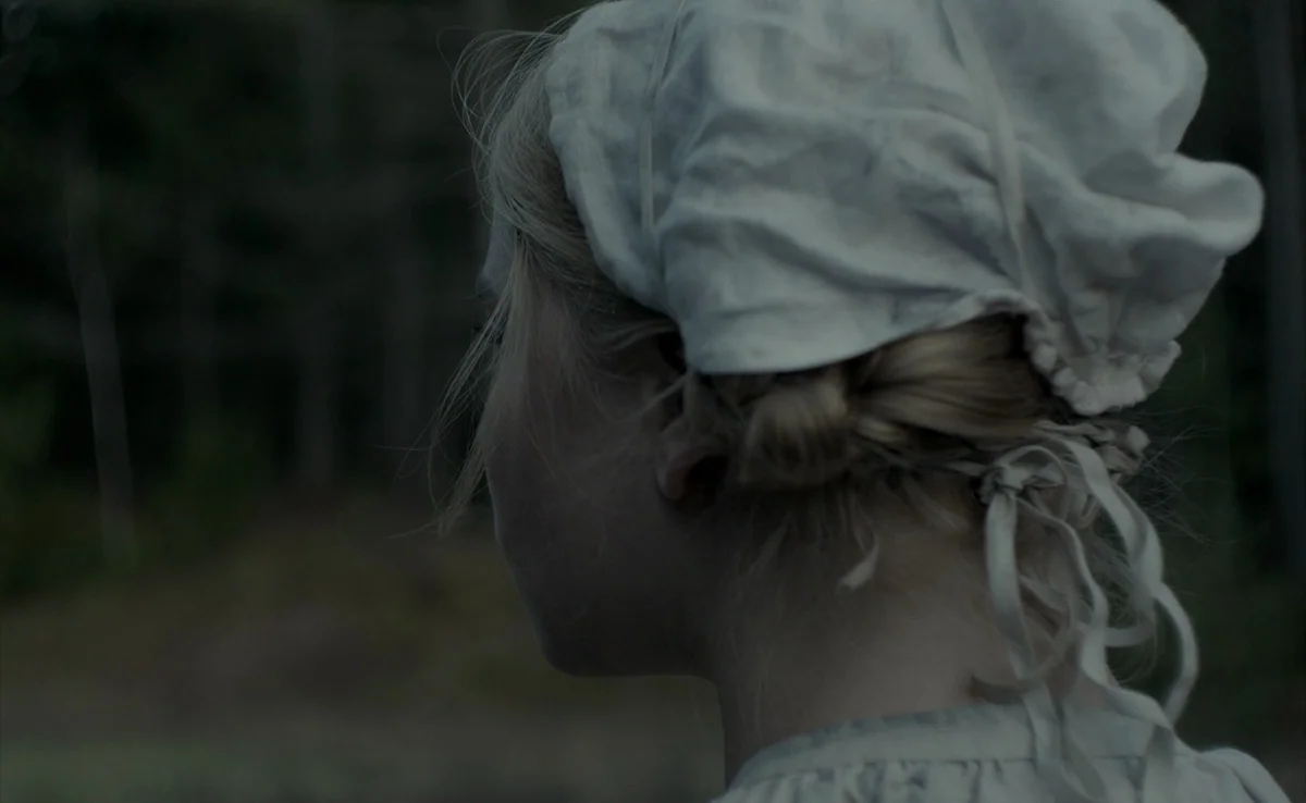 The Witch (2015) movie still: closeup — A dense, dark woodland provides a blurred backdrop behind a young woman. She turns her head to look into the…