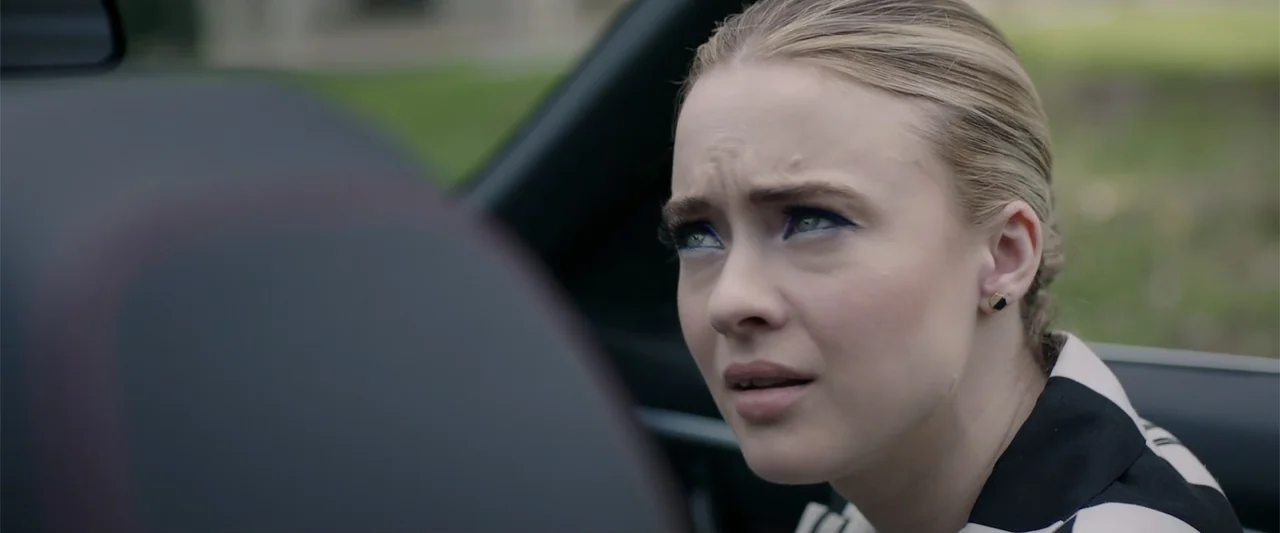 Playing God (2021) movie still: closeup — A close-up shot captures a young woman seated inside a moving vehicle. She glances upward and off-screen…