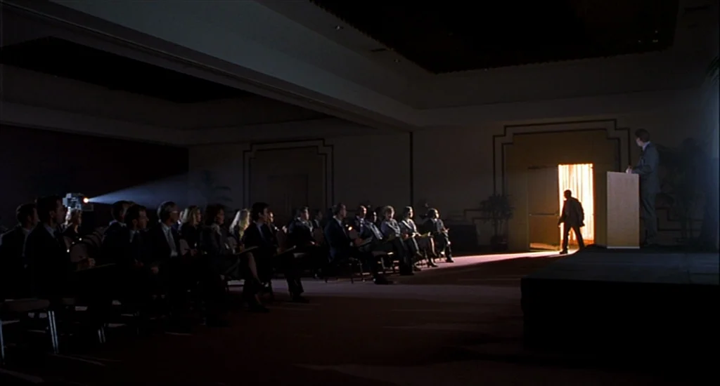 One Hour Photo (2002) movie still: wide — A dark, expansive conference hall with rows of seated attendees facing a lecture podium. A lone man enters…