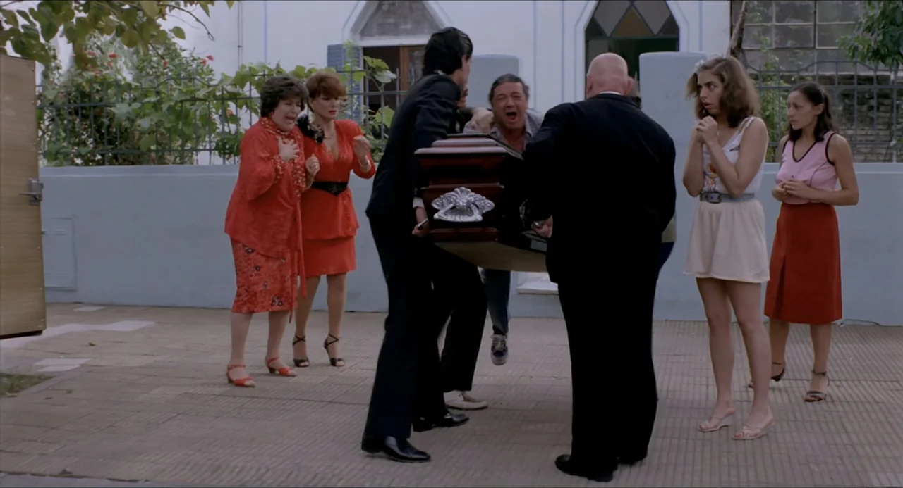 Waiting for the Hearse (1985) movie still: fullbody — A group of people dressed in varying formal and casual attire gather in a paved courtyard.…