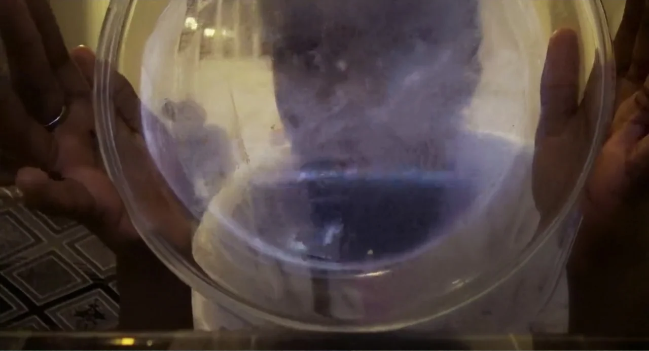 A Dramatic Film (2019) movie still: closeup — An interior space with a person holding a large transparent glass bowl forward. The subject's face is…