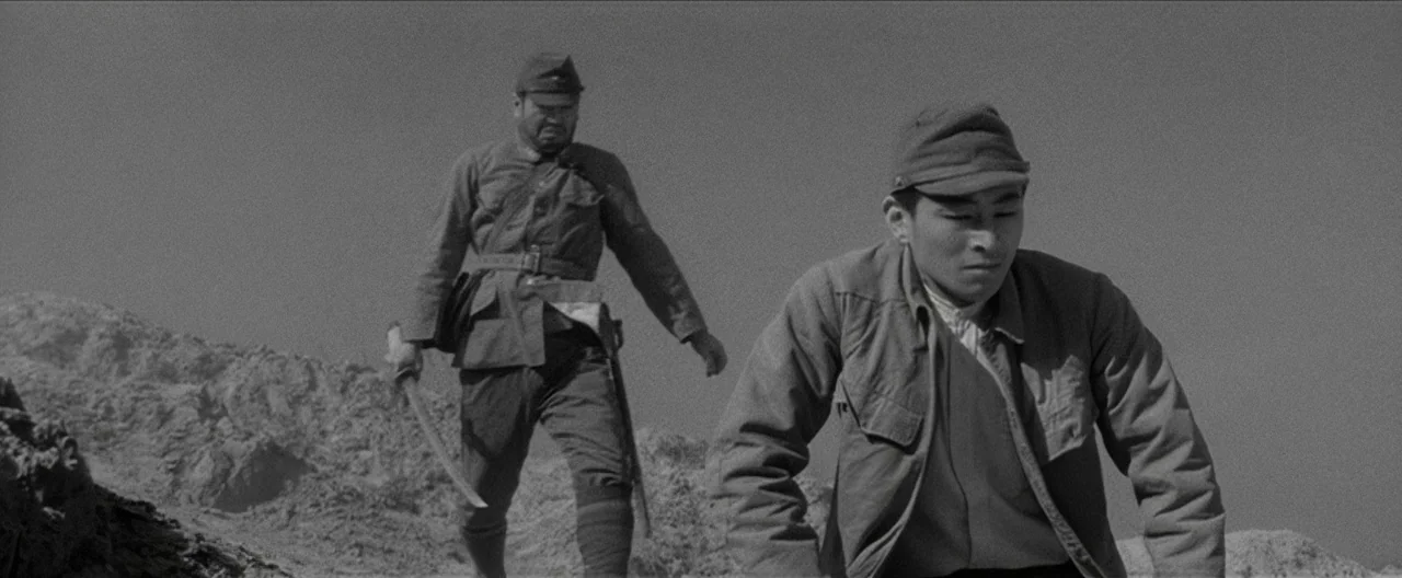 Story of a Prostitute (1965) movie still: fullbody — A desolate rocky desert landscape under a clear, bright sky. Two men walk through the barren…