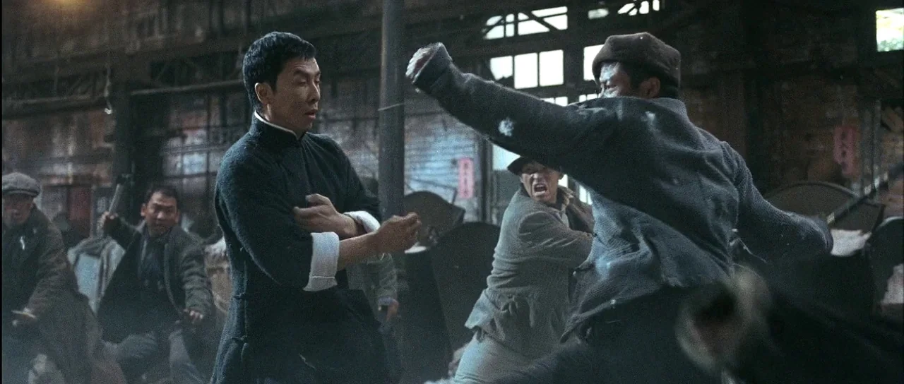 Ip Man (2008) movie still: medium — A dark industrial factory interior features tall windows and exposed brick structural elements. A man in a black…