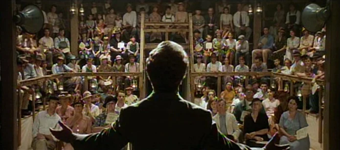 The Neon Bible (1995) movie still: medium — A wooden auditorium is packed with a crowd of people sitting in tiered rows. A man stands in the…