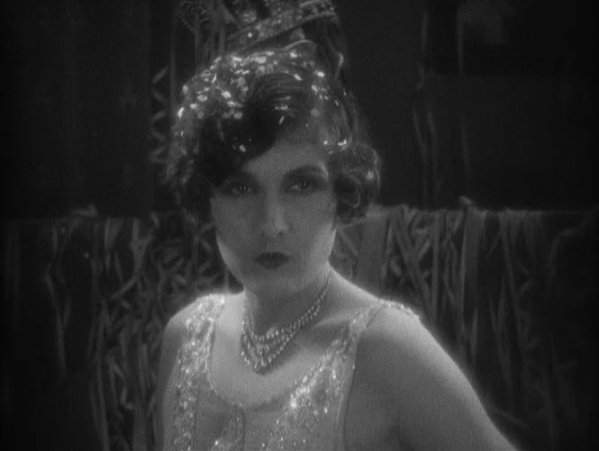 Underworld (1927) movie still: closeup — A festive party setting features a woman adorned with sparkling headwear and pearl necklaces. The woman…