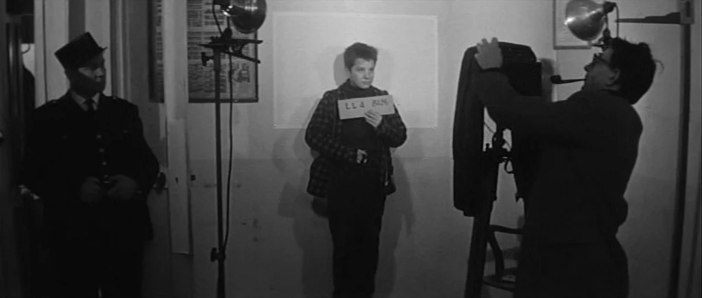 The 400 Blows (1959) movie still: fullbody — A spartan police mugshot room contains a plain white wall and professional lighting equipment. A…