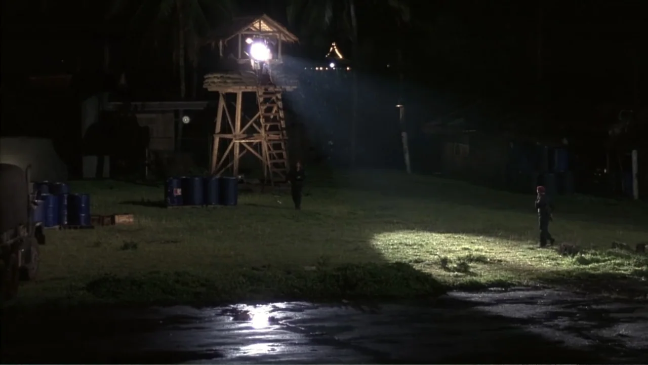 Behind Enemy Lines (2001) movie still: wide — A dark military compound features a wooden watchtower and multiple blue supply barrels. A soldier…