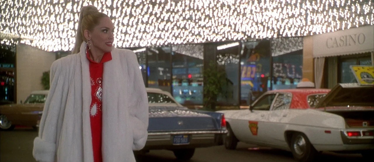 Casino (1995) movie still: medium — A woman stands under a canopy of bright lights outside a building entrance. Vintage cars are parked behind her…
