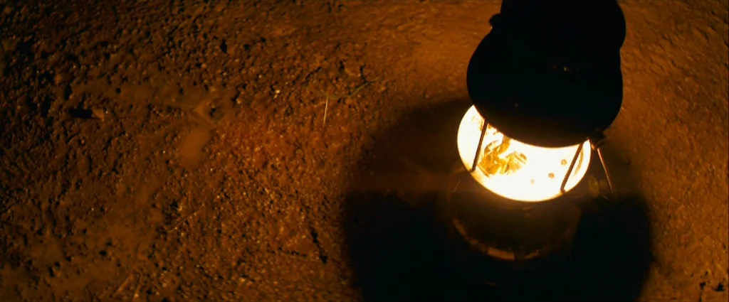 Che Part 1 (The Argentine) (2008) movie still: closeup — A dark, textured patch of dry earth is illuminated by a small lantern. The lantern sits…