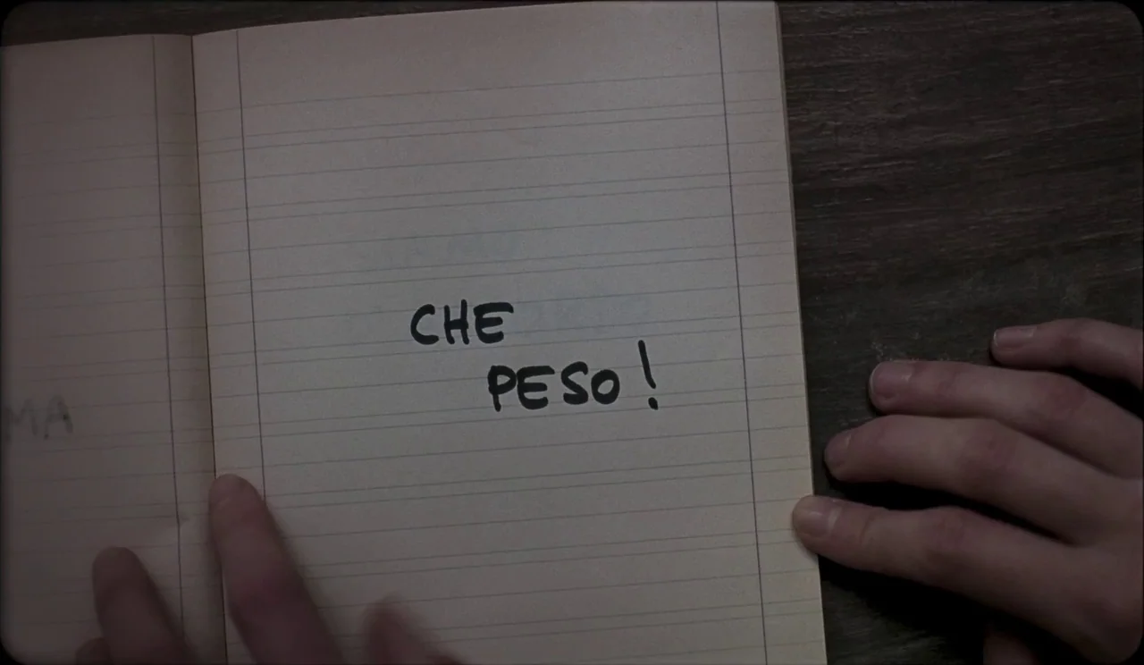 La Chimera (2023) movie still: closeup — An open notebook with lined paper sits on a flat, dark-colored surface. The words CHE PESO! are written in…