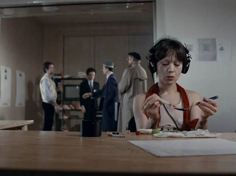 The Third Generation (1979) movie still: medium — A sparse room features a foreground desk and a background area with multiple people. A woman with…