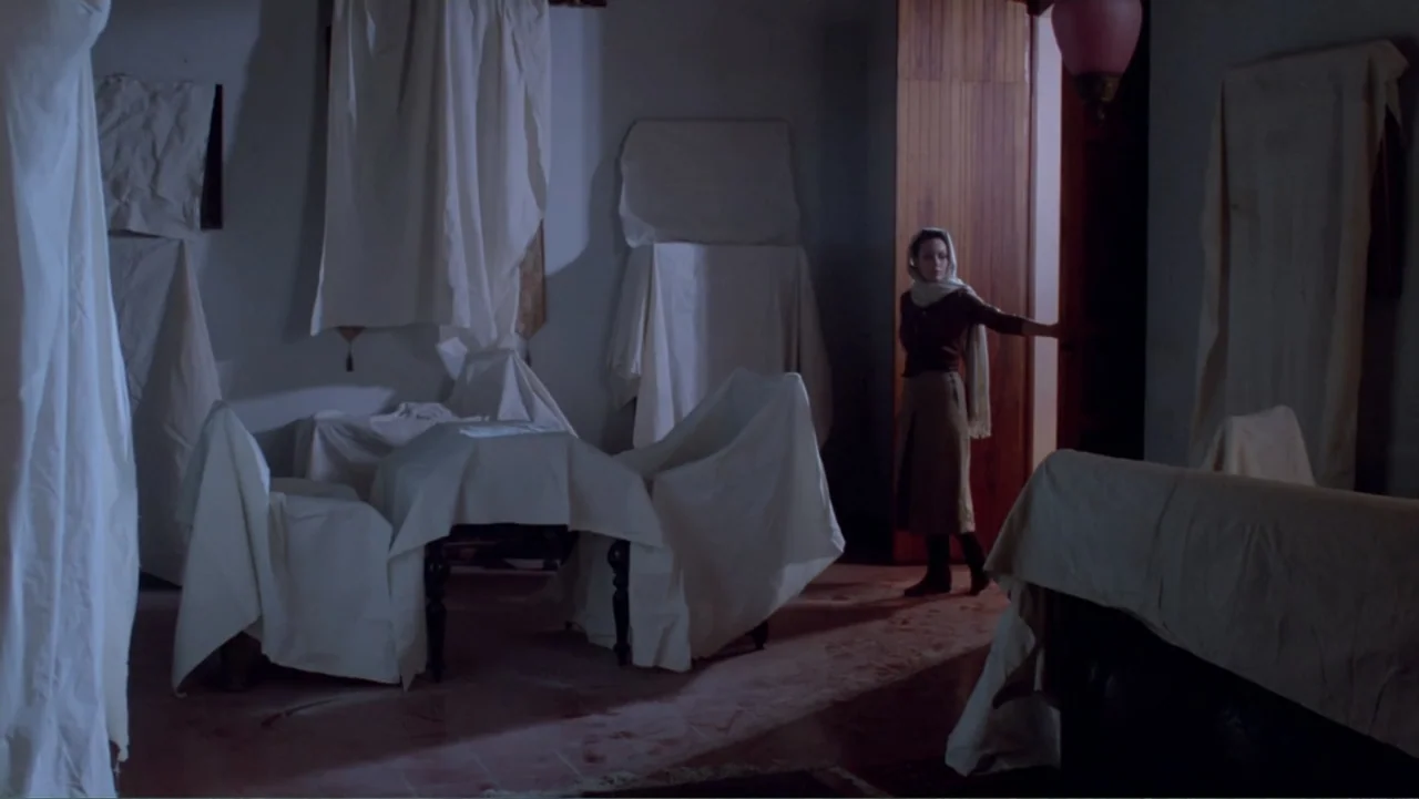 Toña Machetes (1985) movie still: medium — A dimly lit room filled with furniture covered in white sheets. A woman stands in an open doorway while…